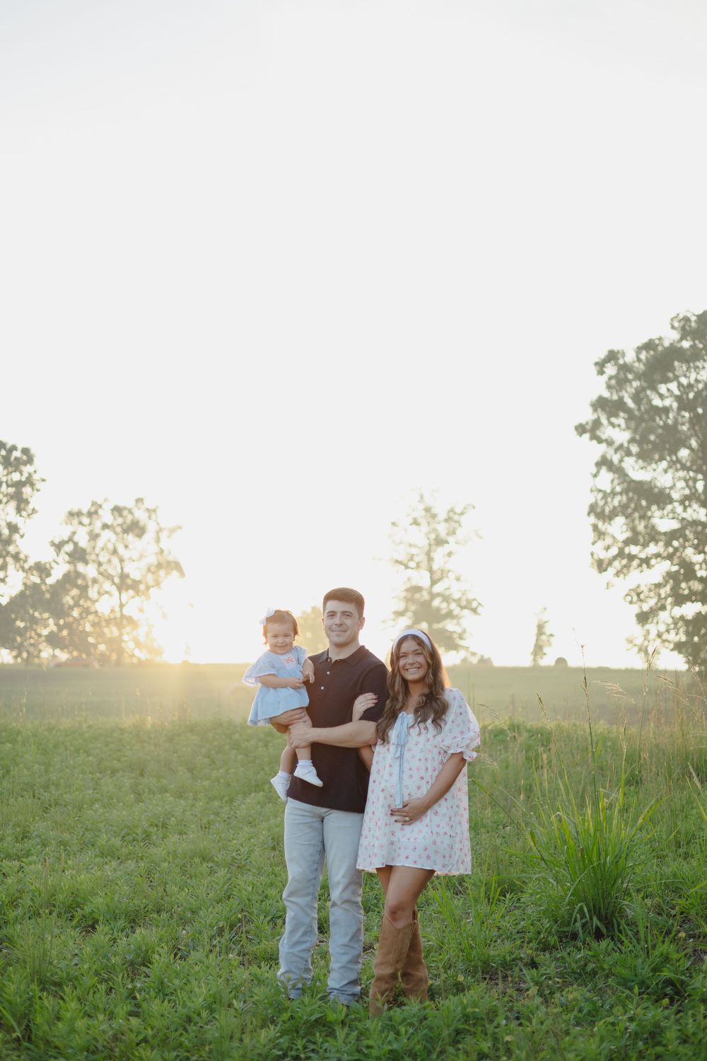 A family stands together in a grassy field during golden hour sunset with trees in the background.