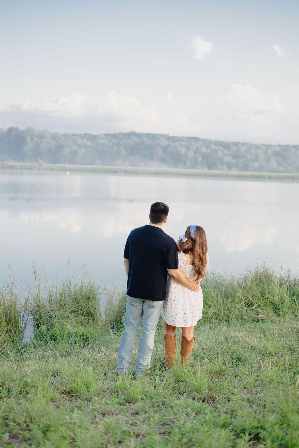A couple stands together on a grassy shoreline overlooking a serene lake on a misty day.