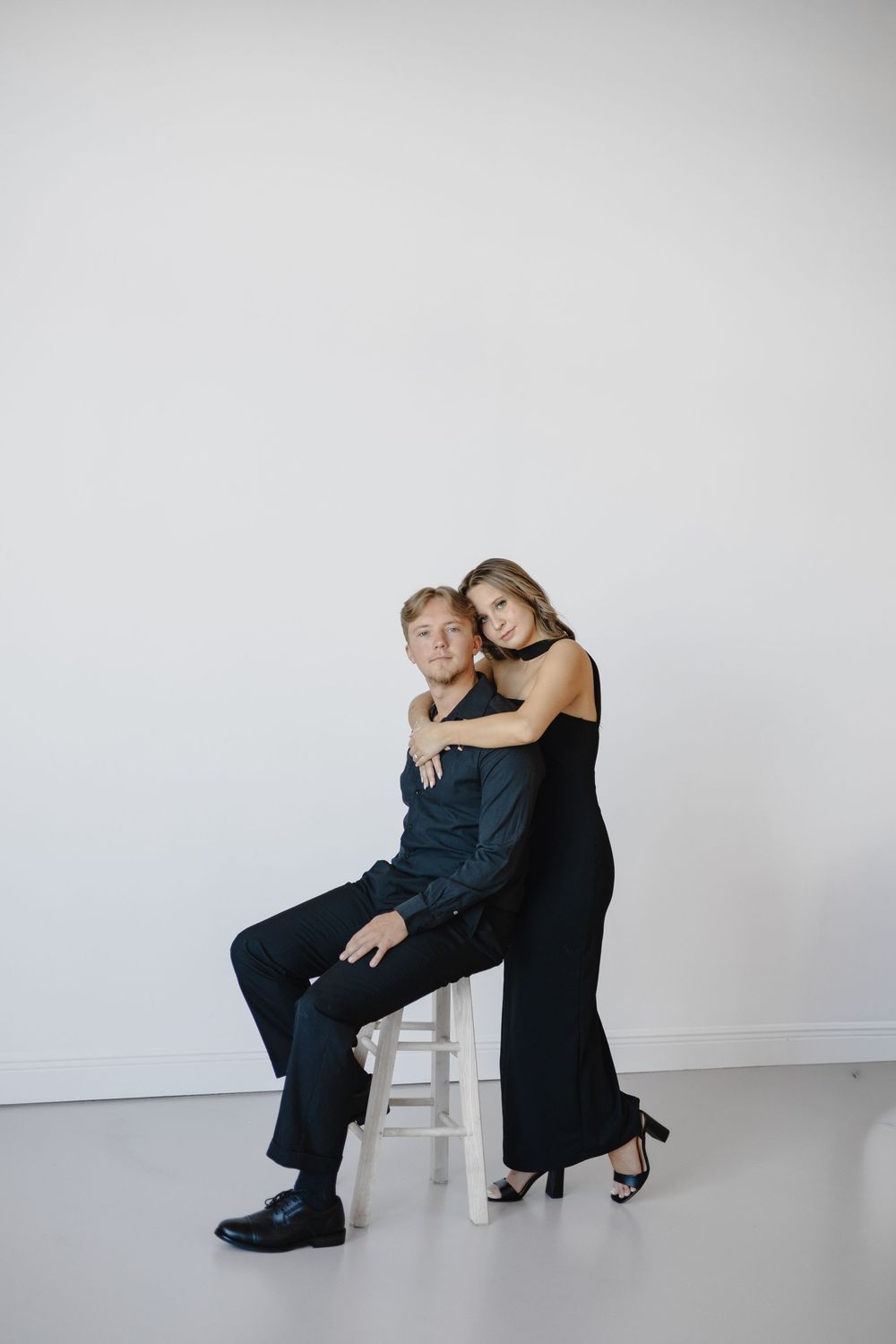 Two people in black attire pose intimately against a white backdrop in a minimalist studio setting.