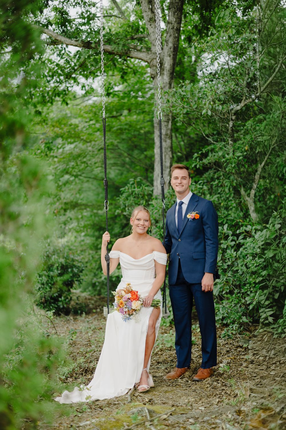 A couple poses together outdoors in wedding attire surrounded by lush green foliage on a sunny day.
