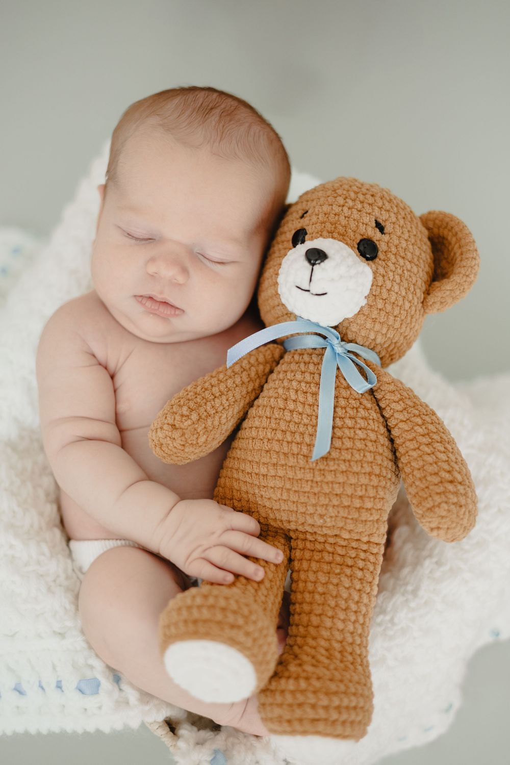 A sleeping baby rests peacefully on white blankets with a crocheted brown teddy bear wearing a blue bow.