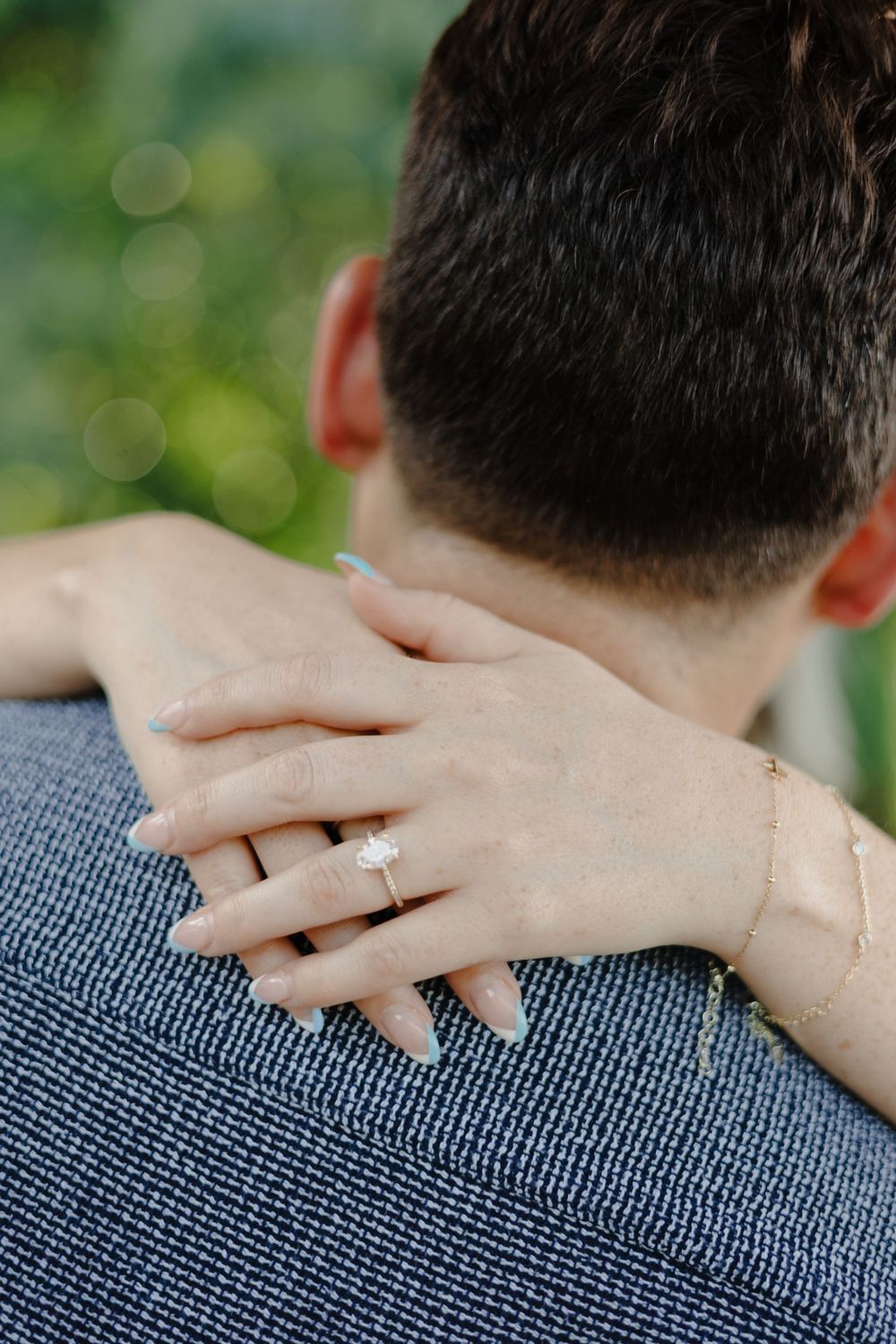 A close-up view of a woman's hand with an engagement ring resting on a man's shoulder wearing a blue shirt.