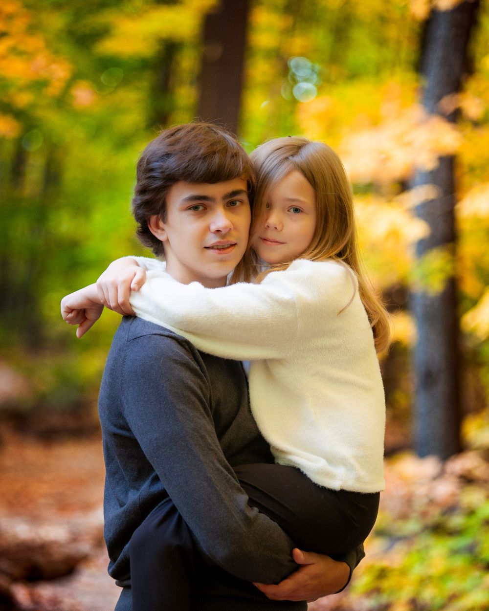 Elder brother and little sister enjoying a joyful moment together in an outdoor fall photoshoot, with the vibrant autumn leaves creating a w