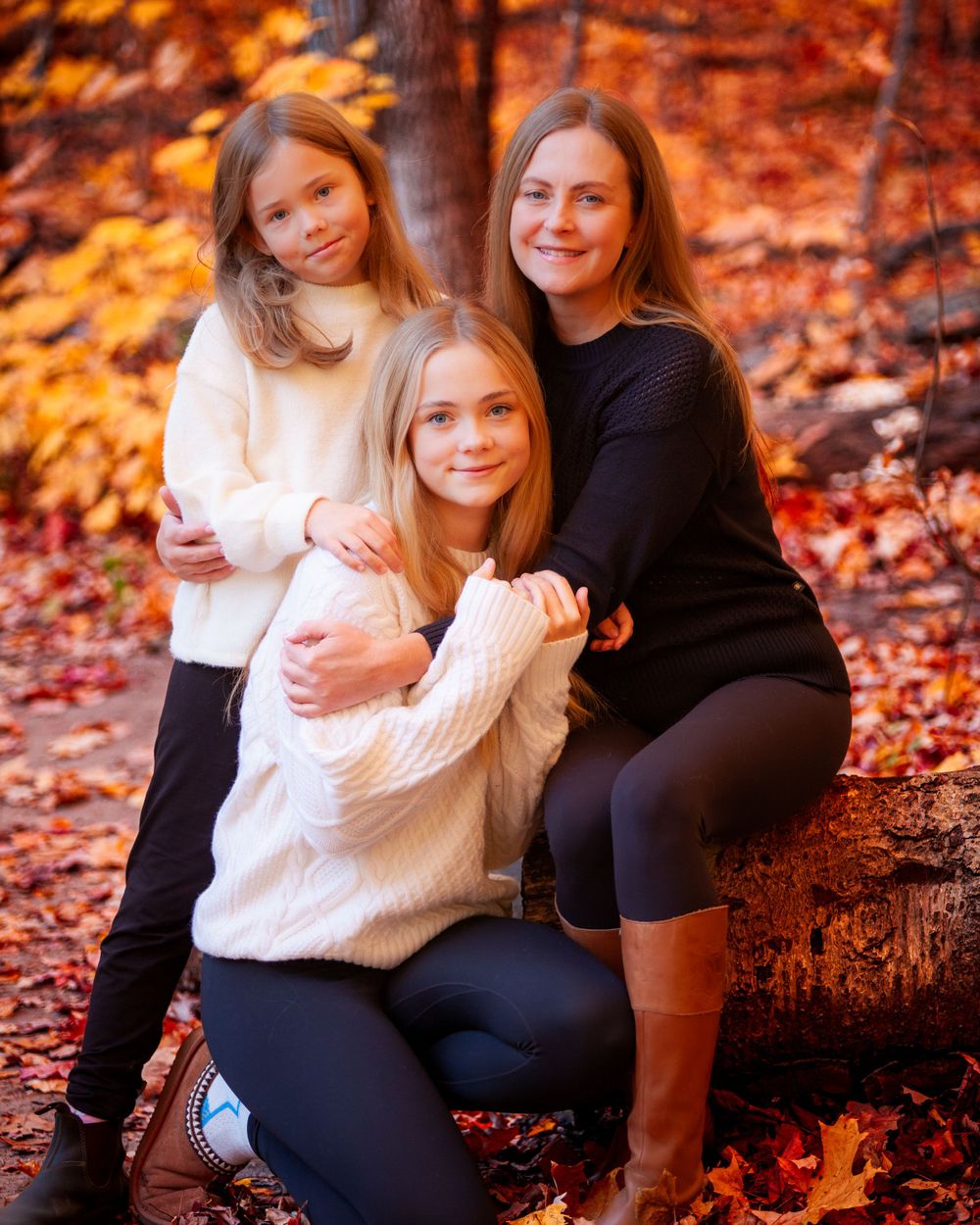 Mother and two sisters—an elder sister and a little sister—posing together in a picturesque outdoor fall setting, with vibrant autumn leaves