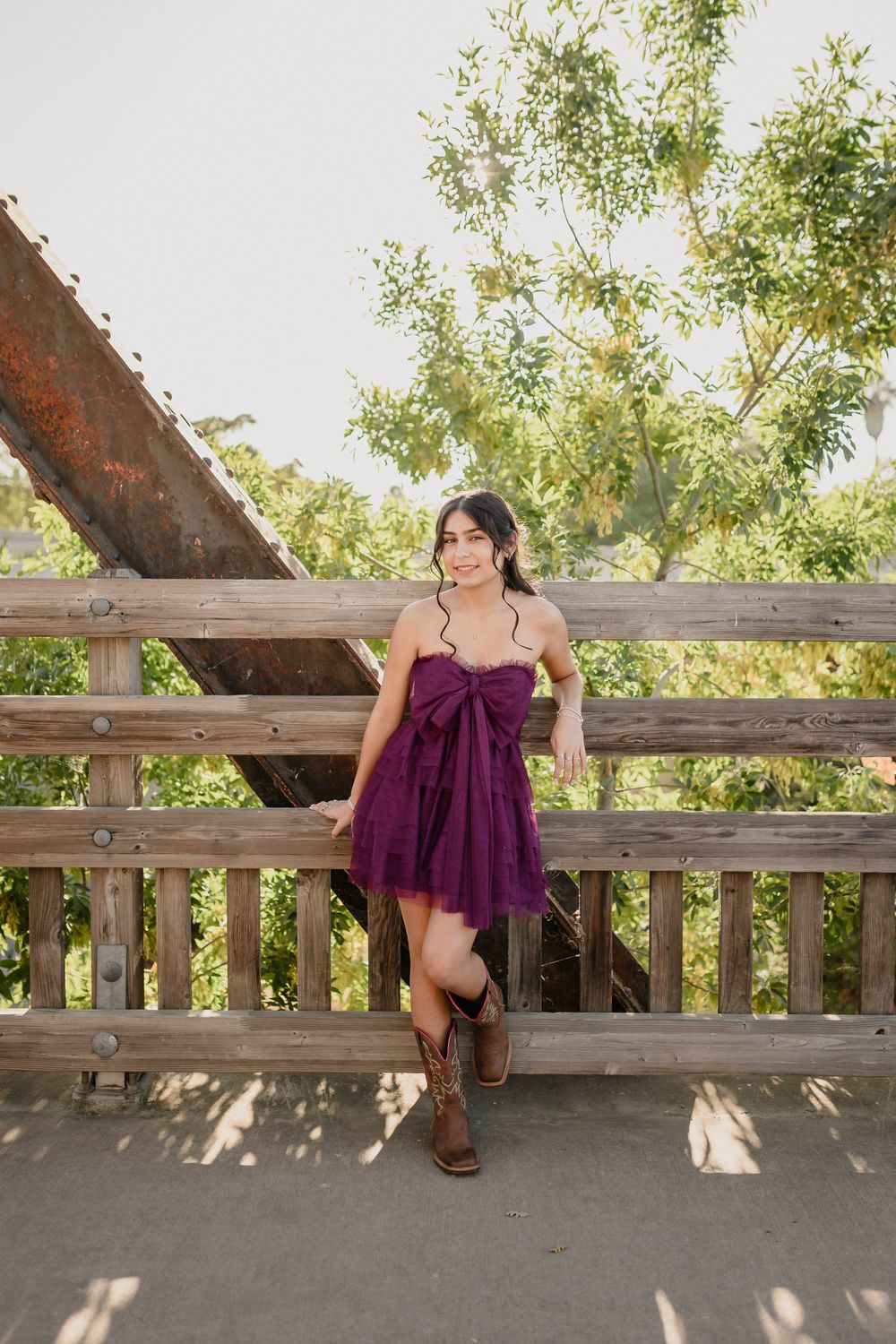 A purple sundress and cowboy boots outfit styled with a rustic wooden bridge backdrop in natural lighting.