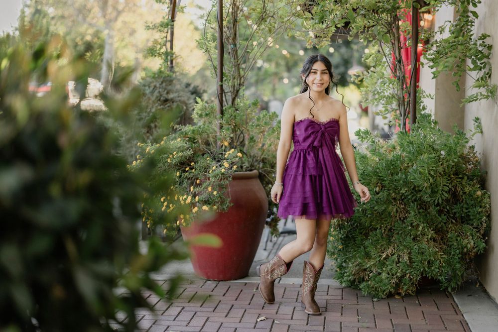 Garden fashion portrait with purple sundress and boots against lush greenery and brick pathway.