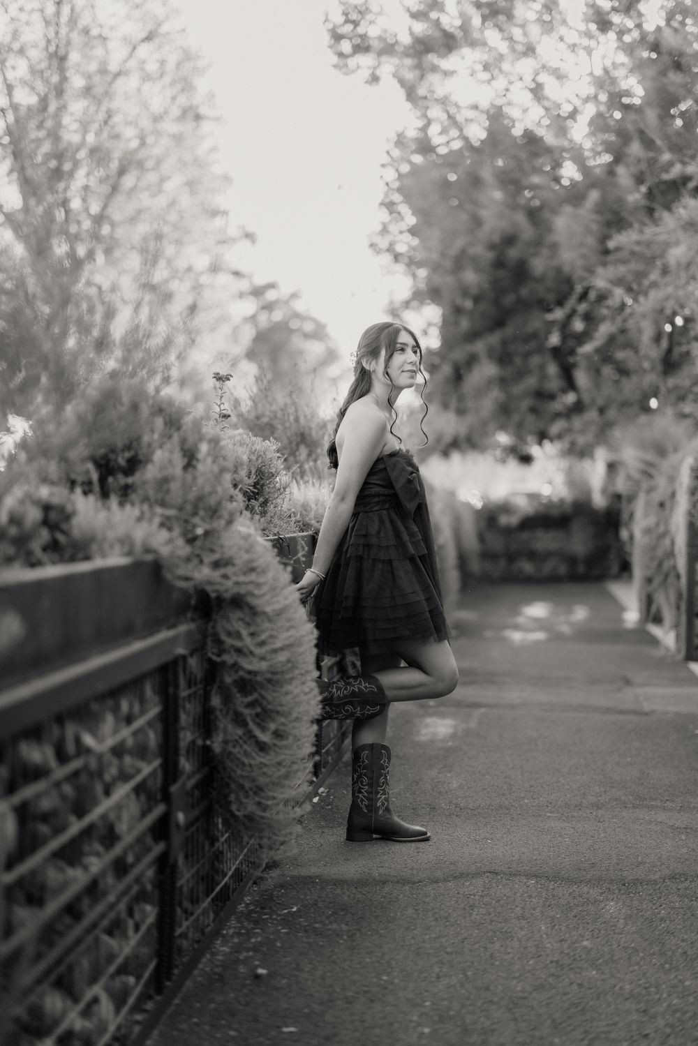 Black and white portrait of fashion model in dress and boots leaning against garden railing.