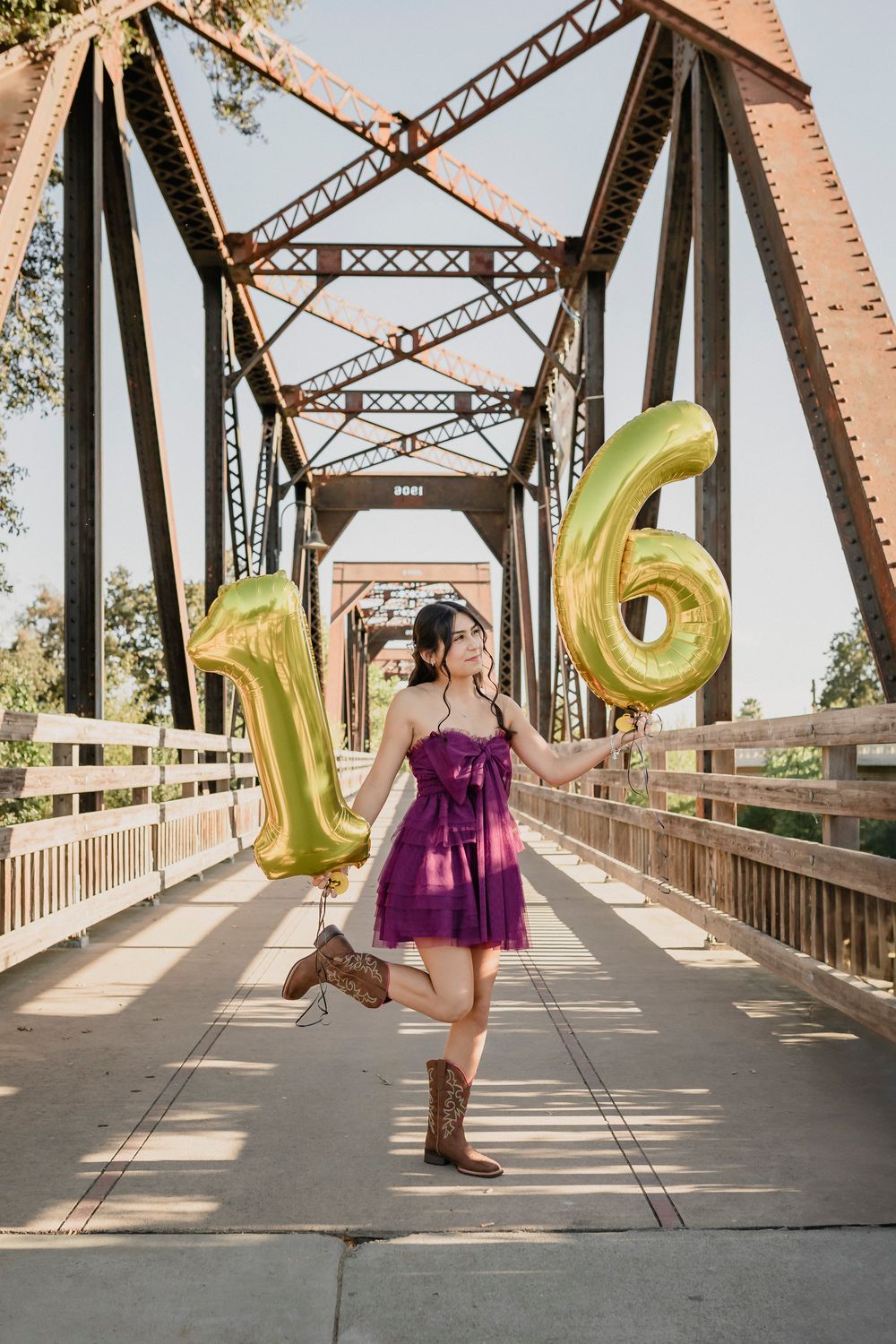 Purple dress and cowboy boots with gold number 16 balloons on an industrial bridge.