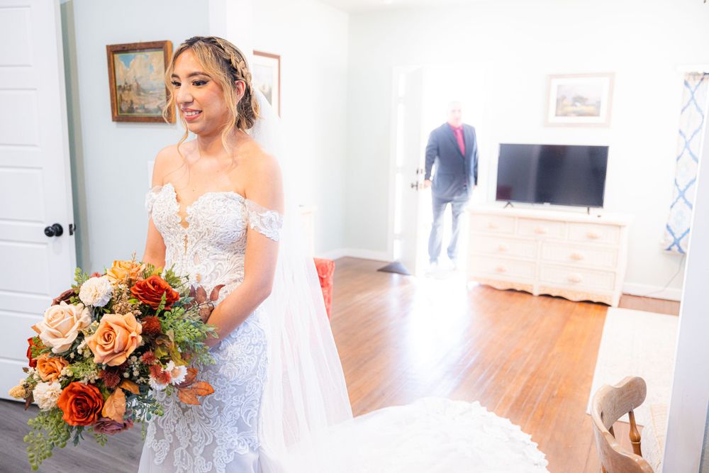 A bride in a white lace gown holds a vibrant autumn-colored bouquet while standing in a bright room.