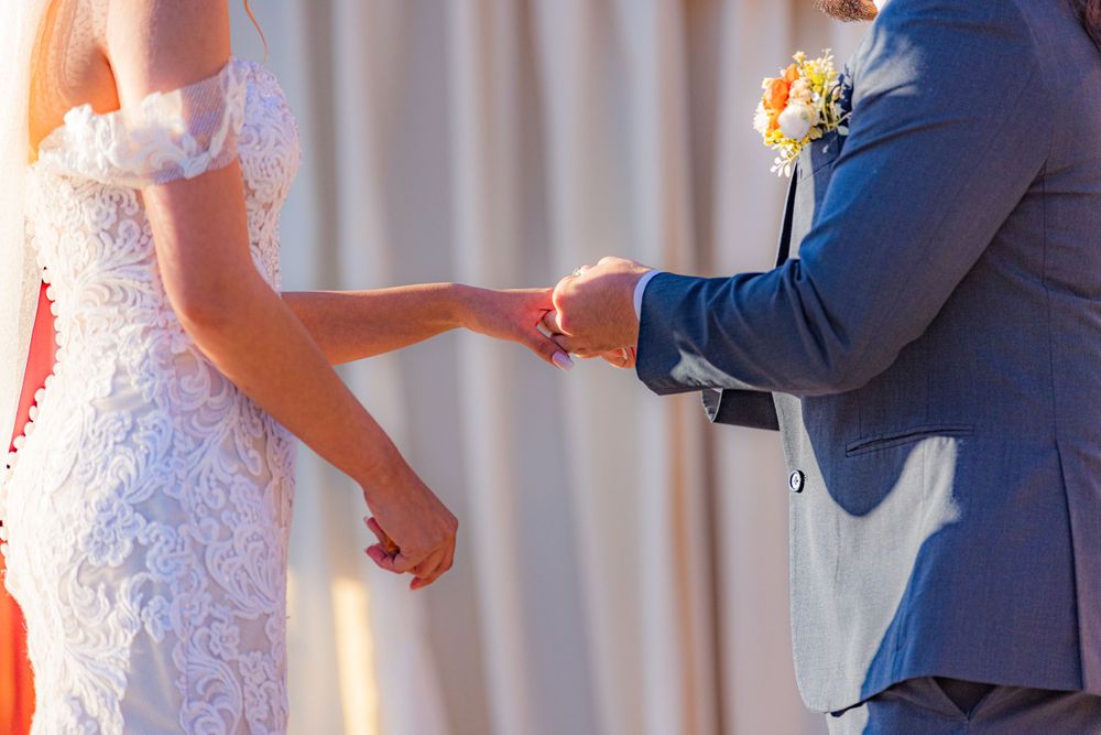 Close up view of a couple holding hands during wedding ceremony with bride in lace dress and groom in blue suit.