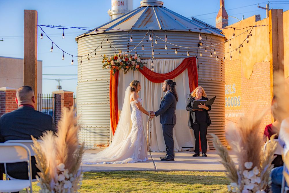 A wedding ceremony takes place in a rustic silo venue decorated with string lights, red drapes and pampas grass at sunset.