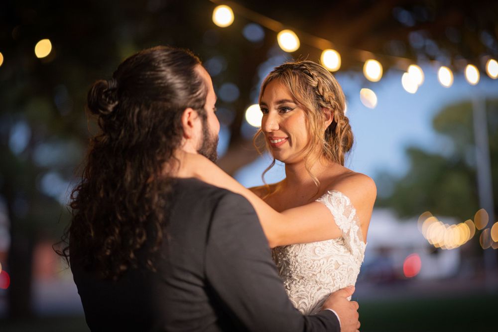 A couple dances together at an outdoor evening wedding reception with glowing string lights in the background.