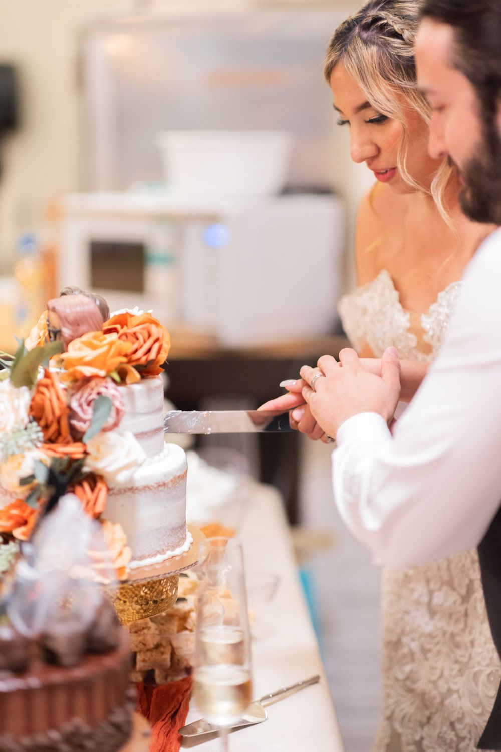 A couple cuts their wedding cake adorned with orange roses and white frosting at their reception celebration.
