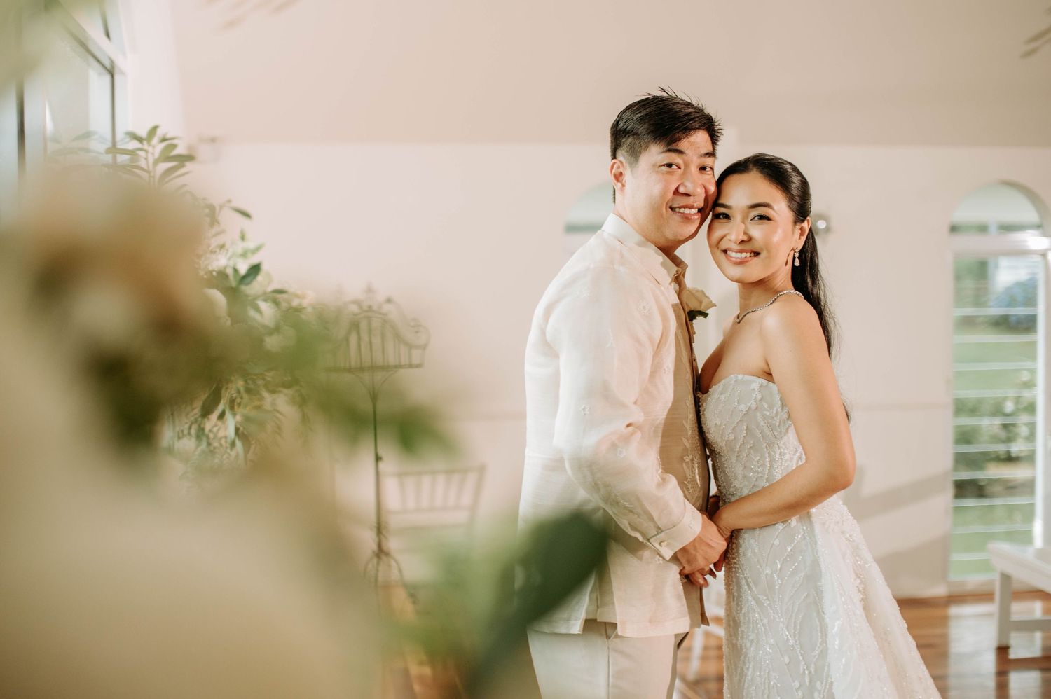 A couple shares a tender moment in a sunlit room wearing elegant white wedding attire.