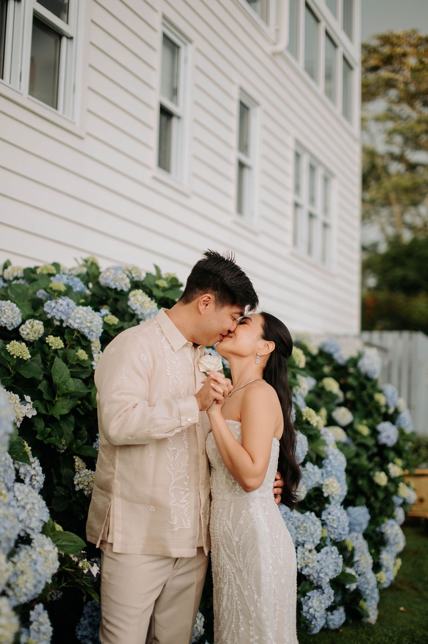Romantic moment by white hydrangea bushes against white building exterior.
