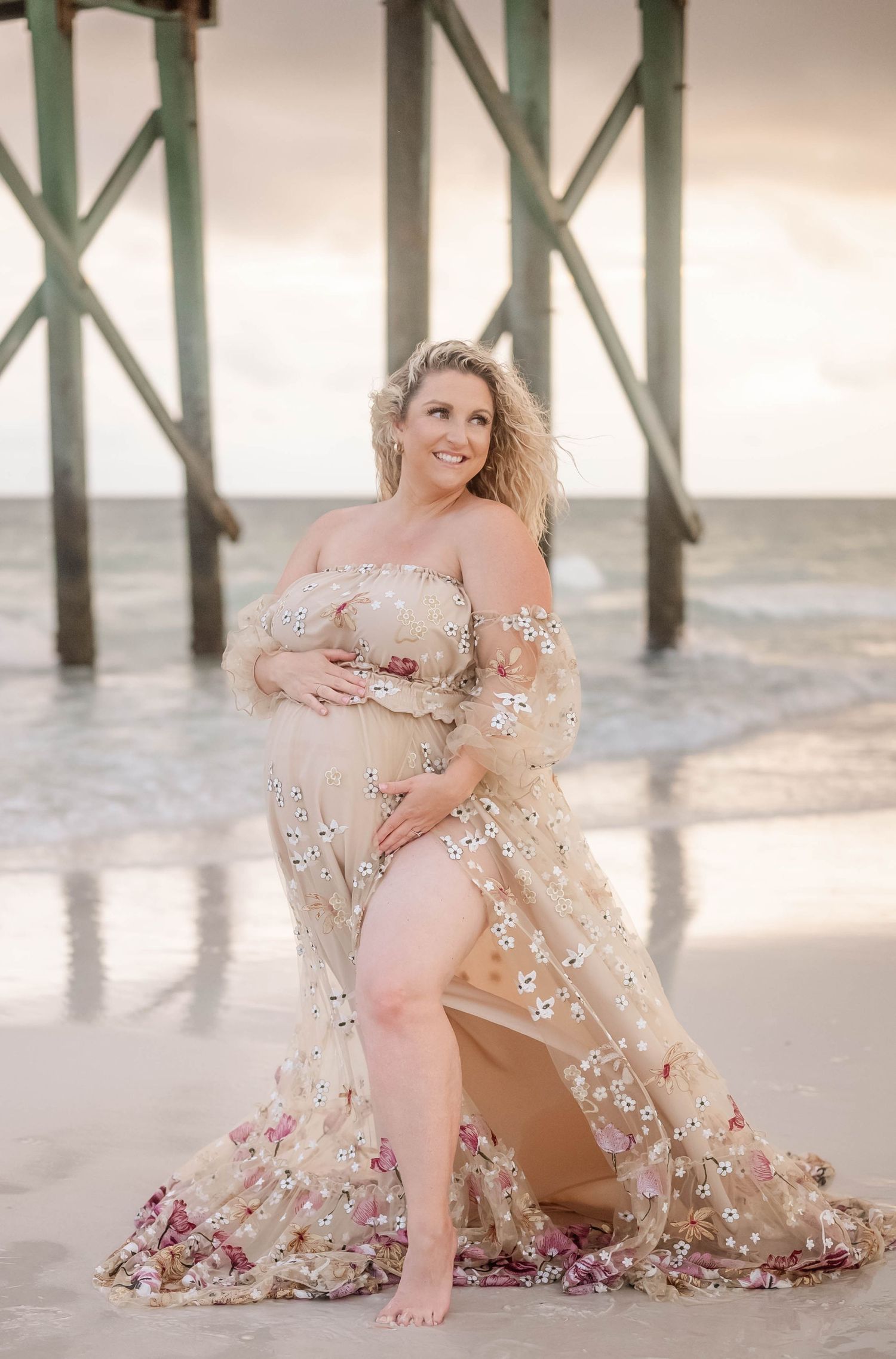 Elegant maternity beach photo with flowing champagne dress adorned with floral details near pier.