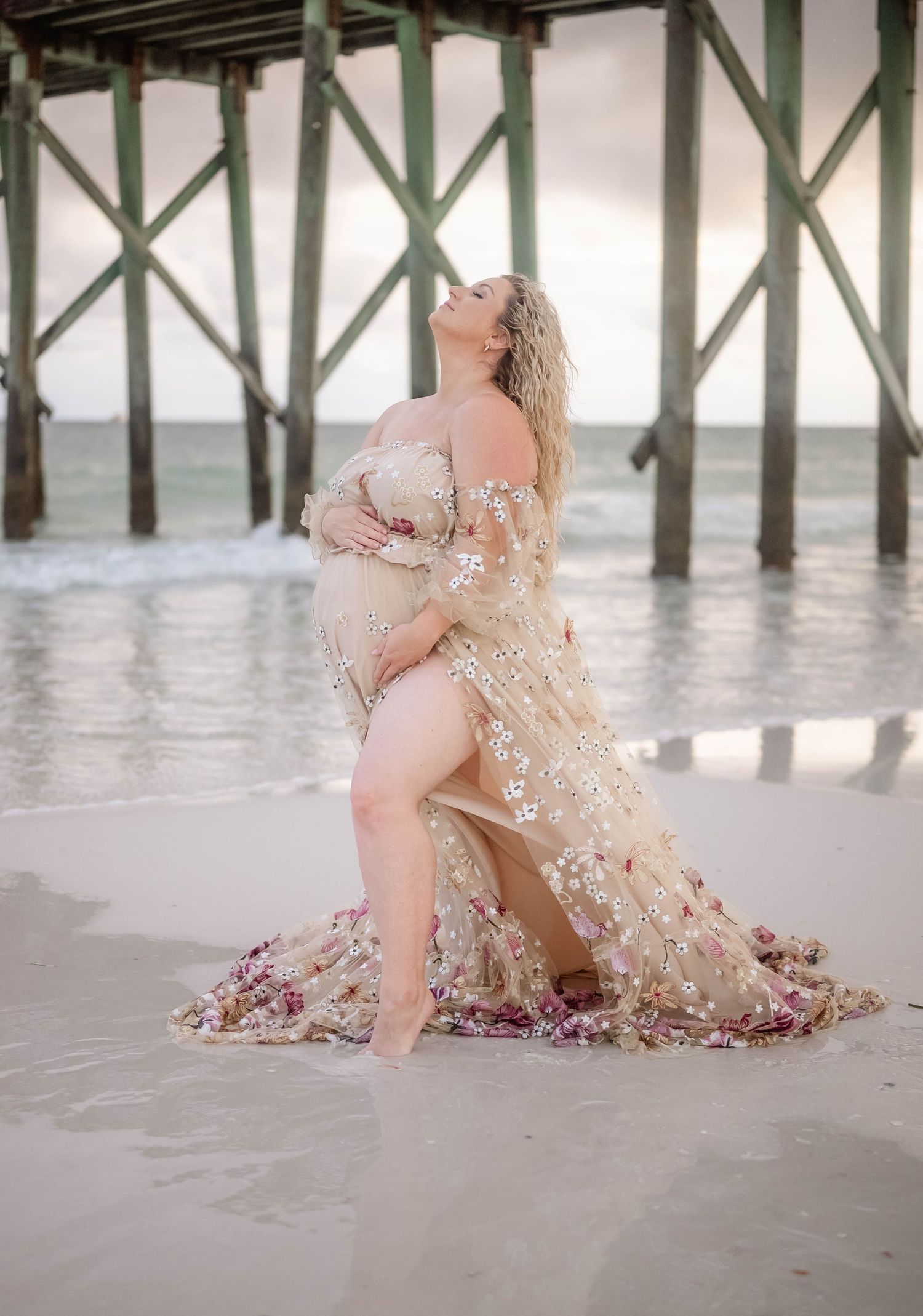 Beach maternity pose with sparkly dress near wooden pier at sunset.