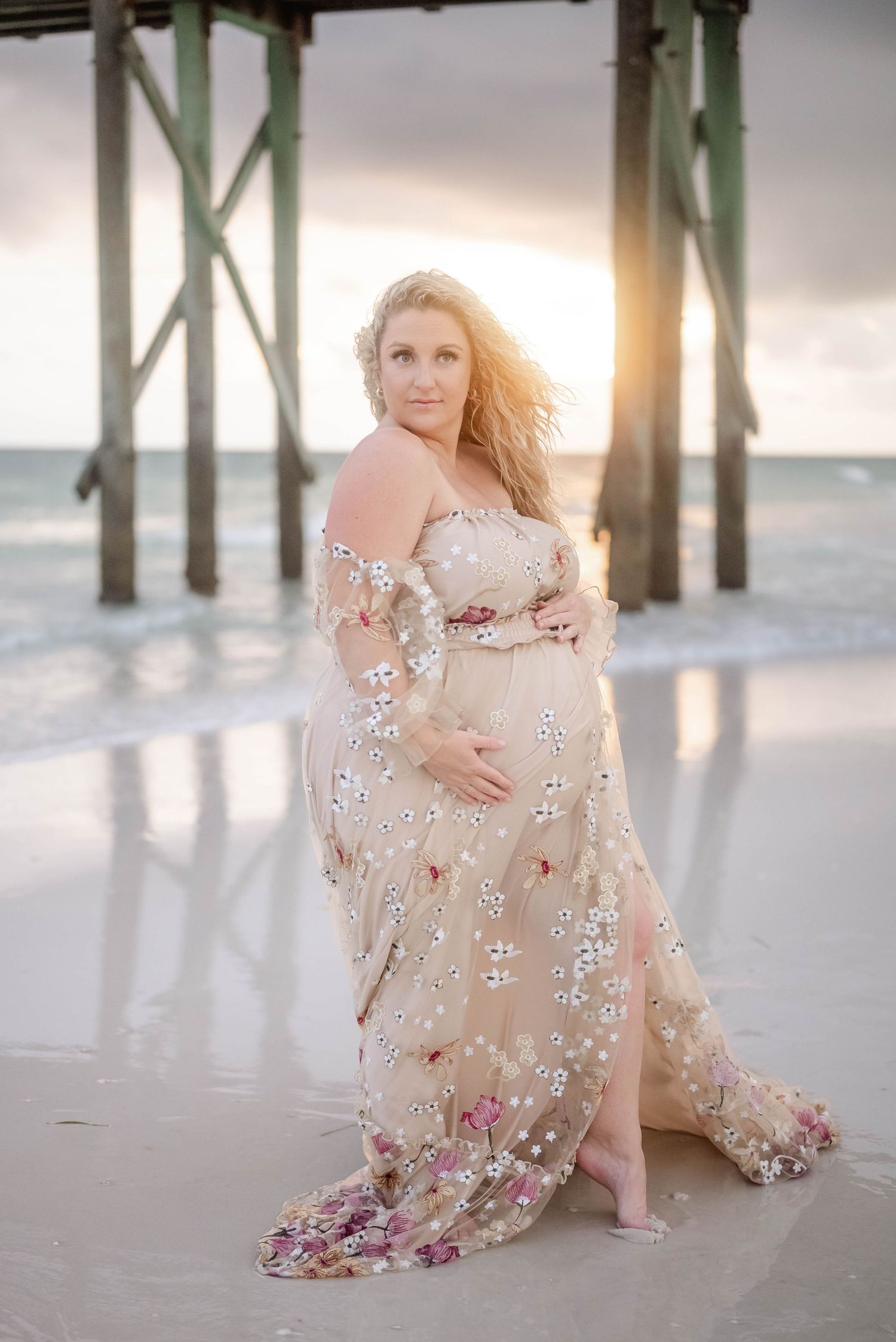 Beach maternity portrait with flowing floral dress and wooden pier backdrop.