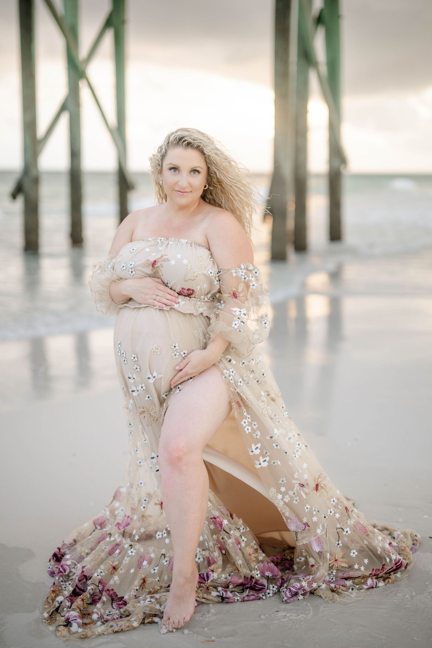 Elegant maternity portrait on beach with detailed floral dress and pier backdrop.