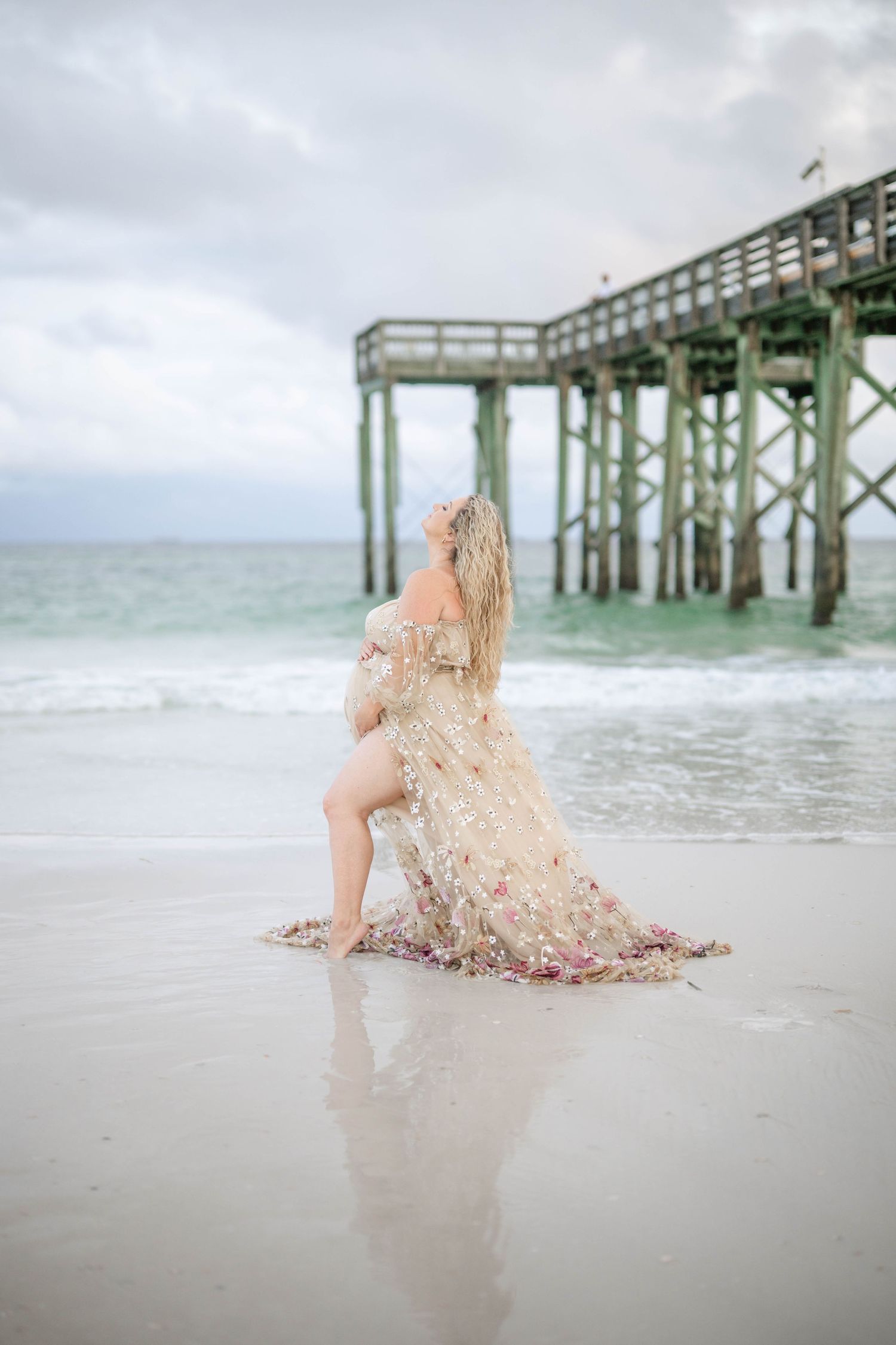 Dreamy beach maternity photo with sheer floral dress near wooden pier.