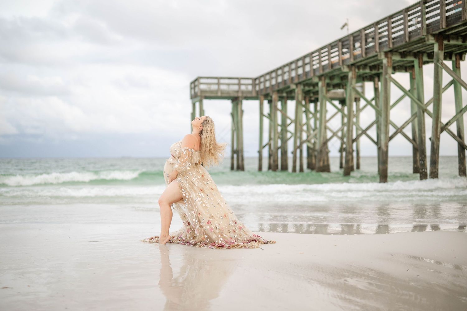 Artistic maternity beach photo with flowing dress near oceanside pier structure.