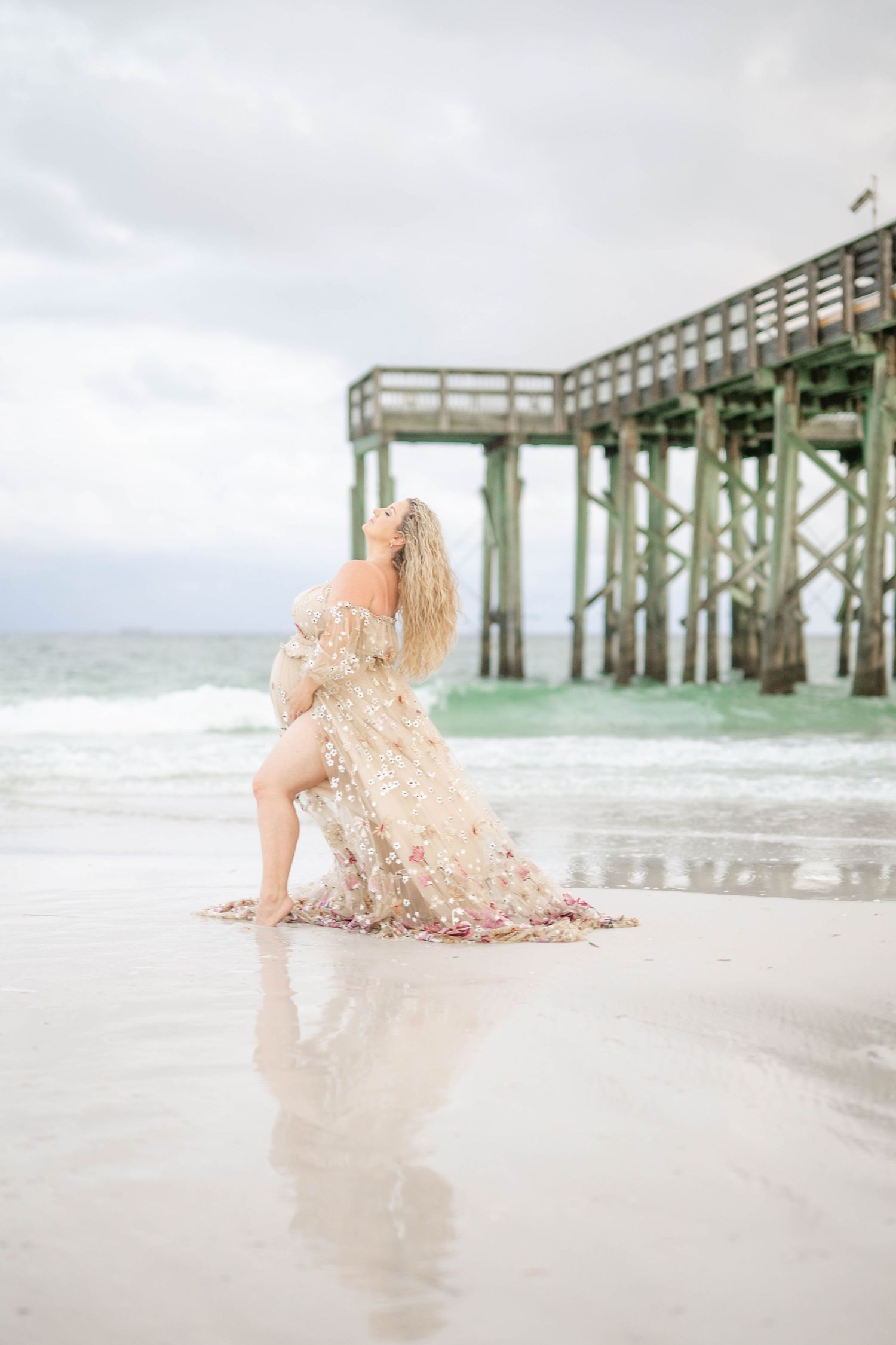 Ethereal maternity photo of woman in champagne gown posing on beach near wooden pier during sunset.