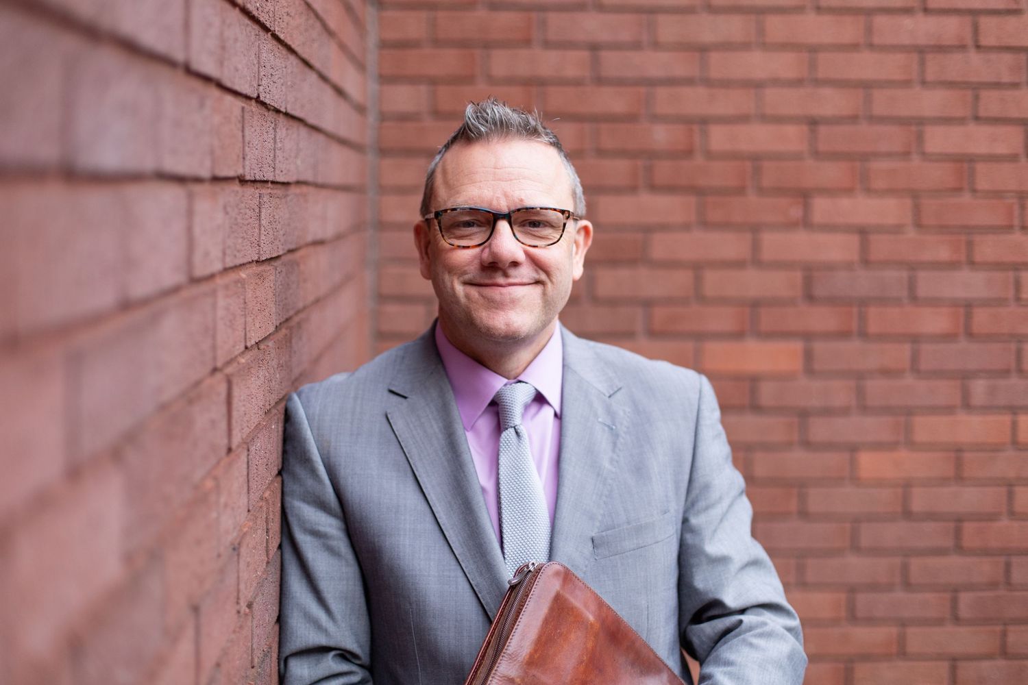 Professional in a gray suit and purple tie smiling while standing against a brick wall background.