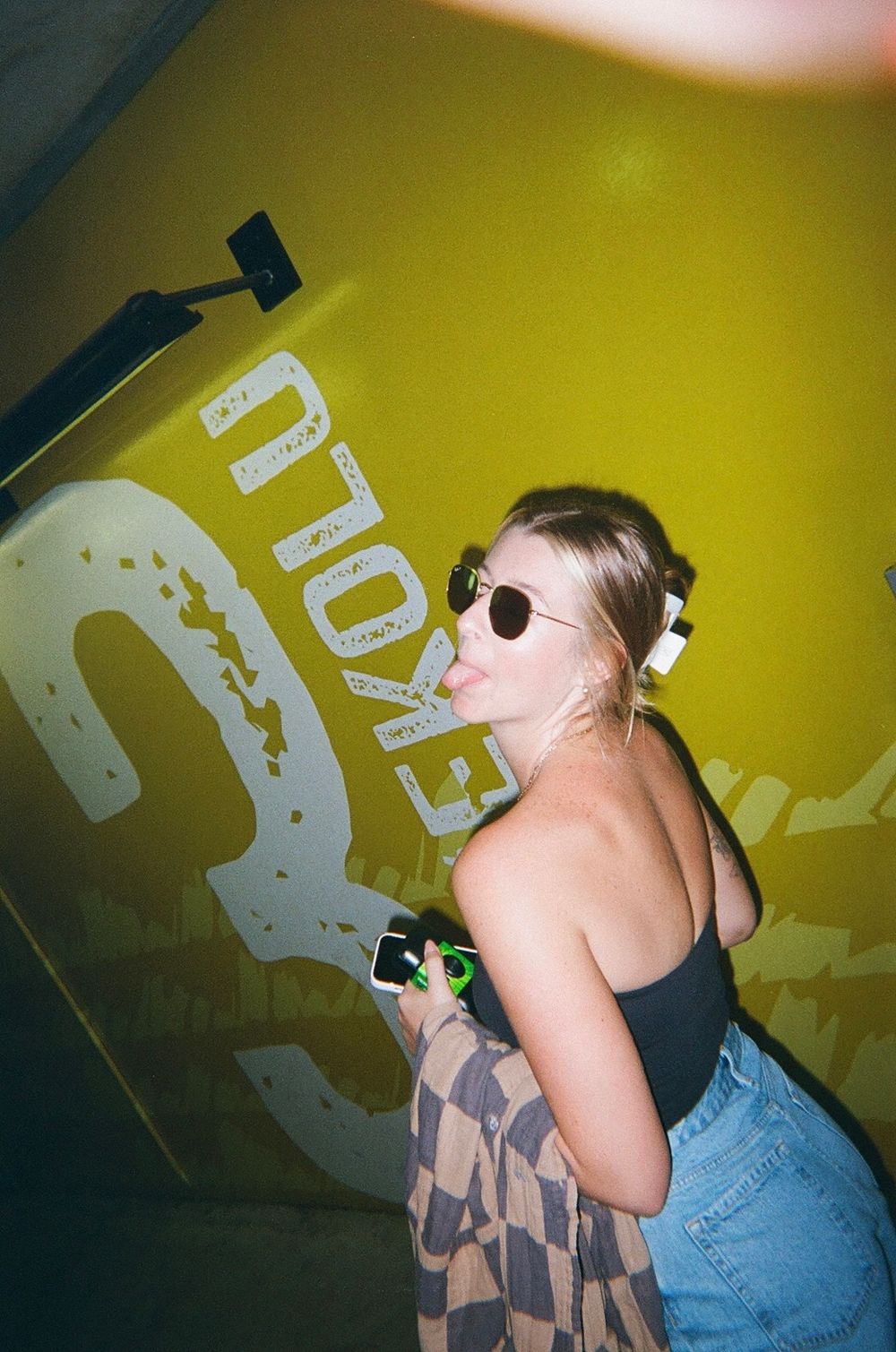 Person in black top and jeans with sunglasses standing against yellow wall with text.