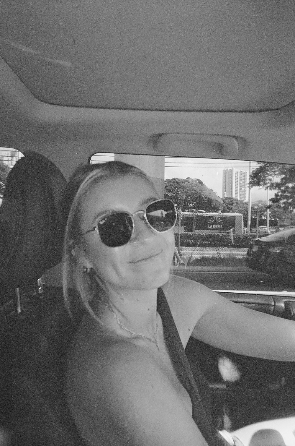 Person wearing sunglasses taking selfie from car driver's seat on sunny day.