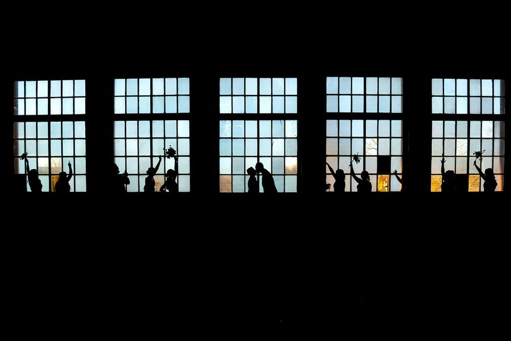 A row of bright windows illuminates a dark industrial building interior with silhouetted figures visible.