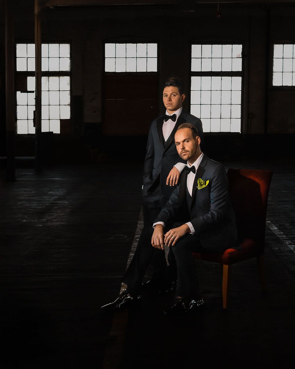 Two people in black tuxedos and bow ties pose in a moody industrial space with large windows.