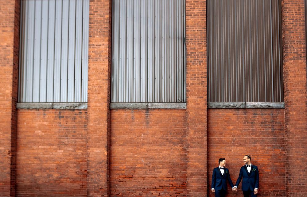 Two people in business attire stand in front of a red brick industrial building with tall windows.