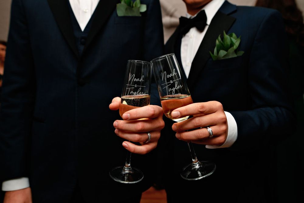 Two people in dark suits with green boutonnieres toast with champagne flutes in an elegant celebration.