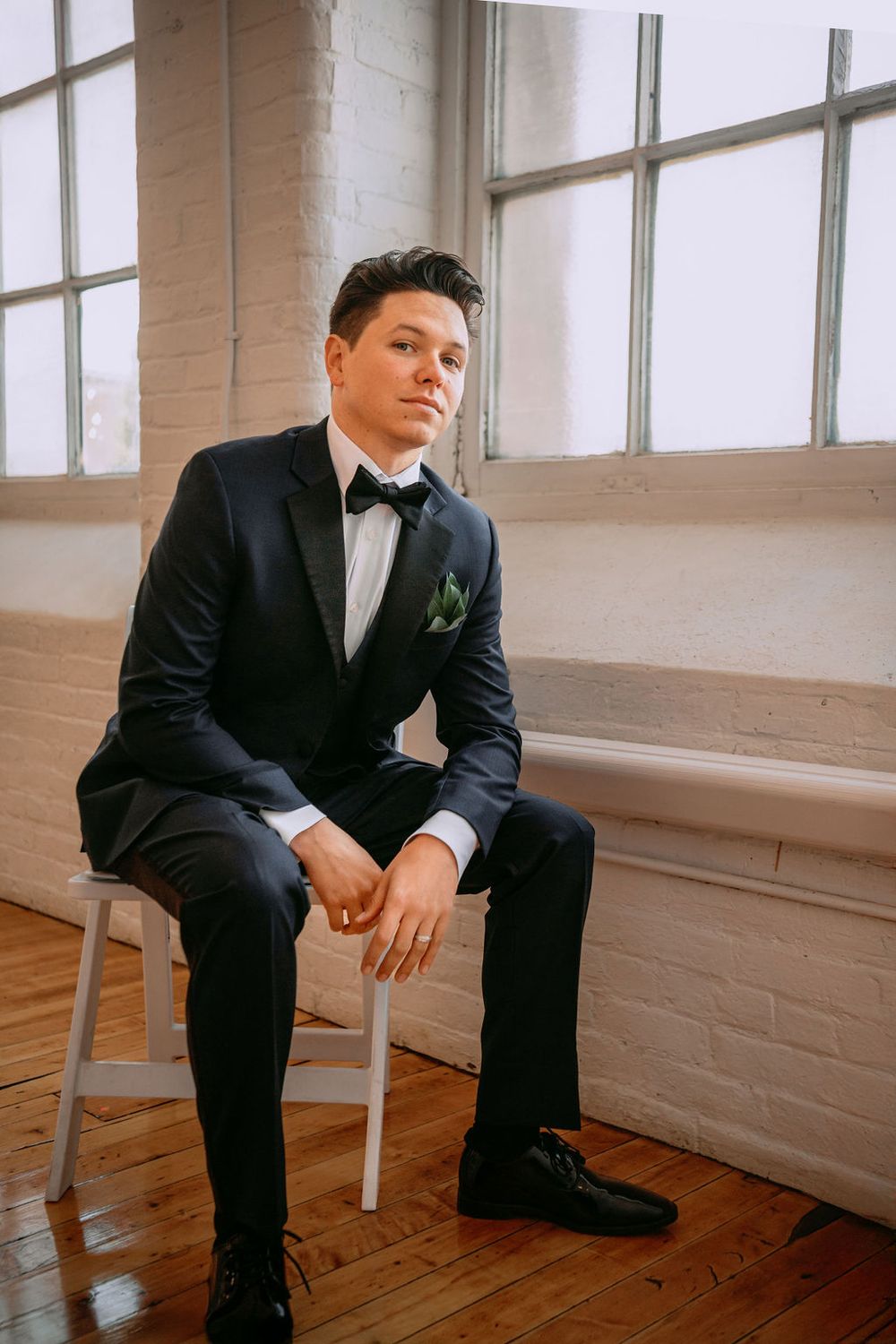 A formal portrait of someone in a classic black tuxedo with green boutonniere seated by a window.