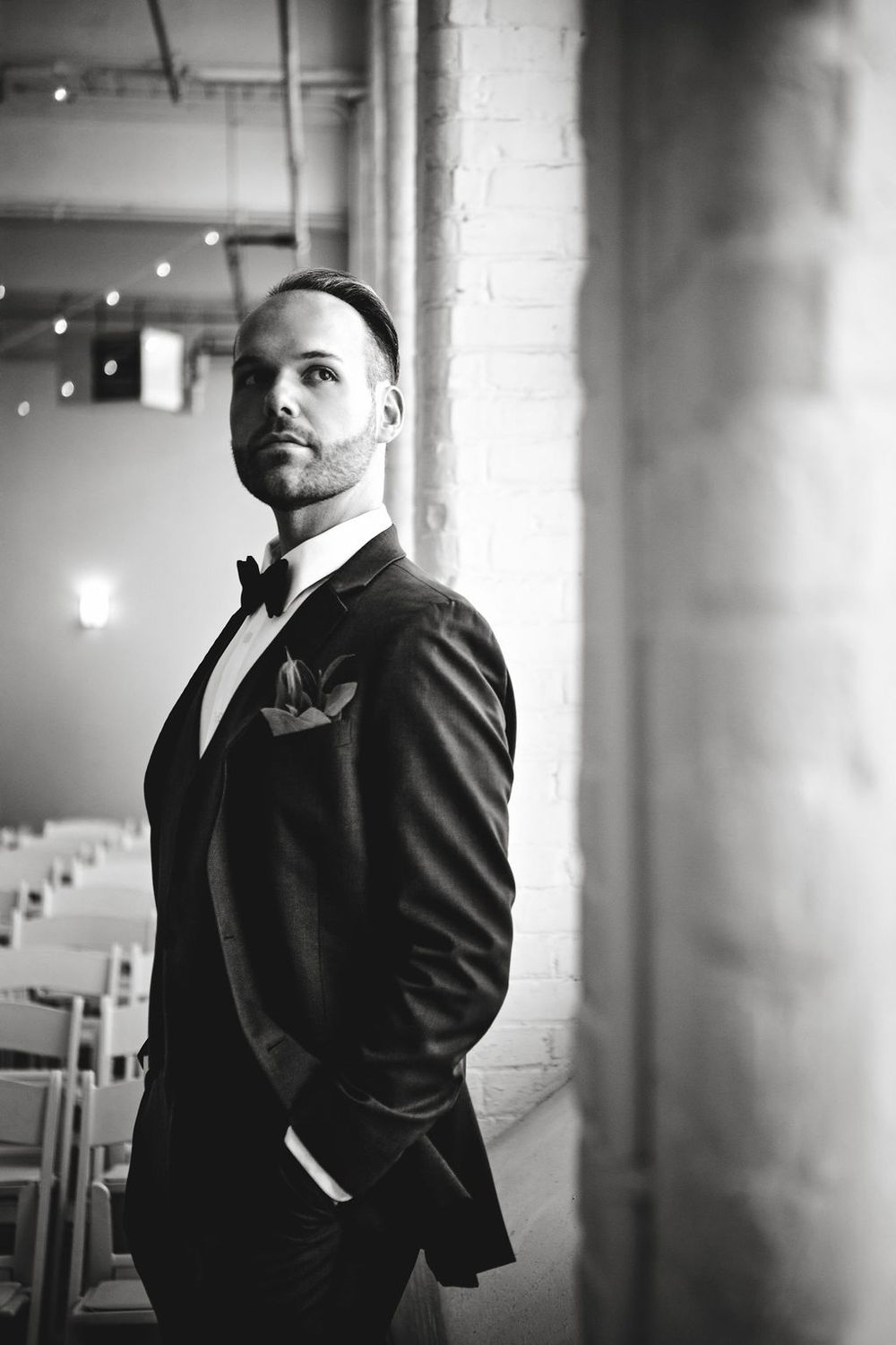 A black and white portrait of someone in a formal tuxedo with bow tie standing in an elegant venue with string lights.
