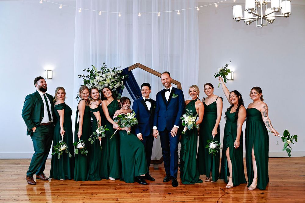 Wedding party poses together in formal attire with bridesmaids in emerald green dresses and groomsmen in dark suits.