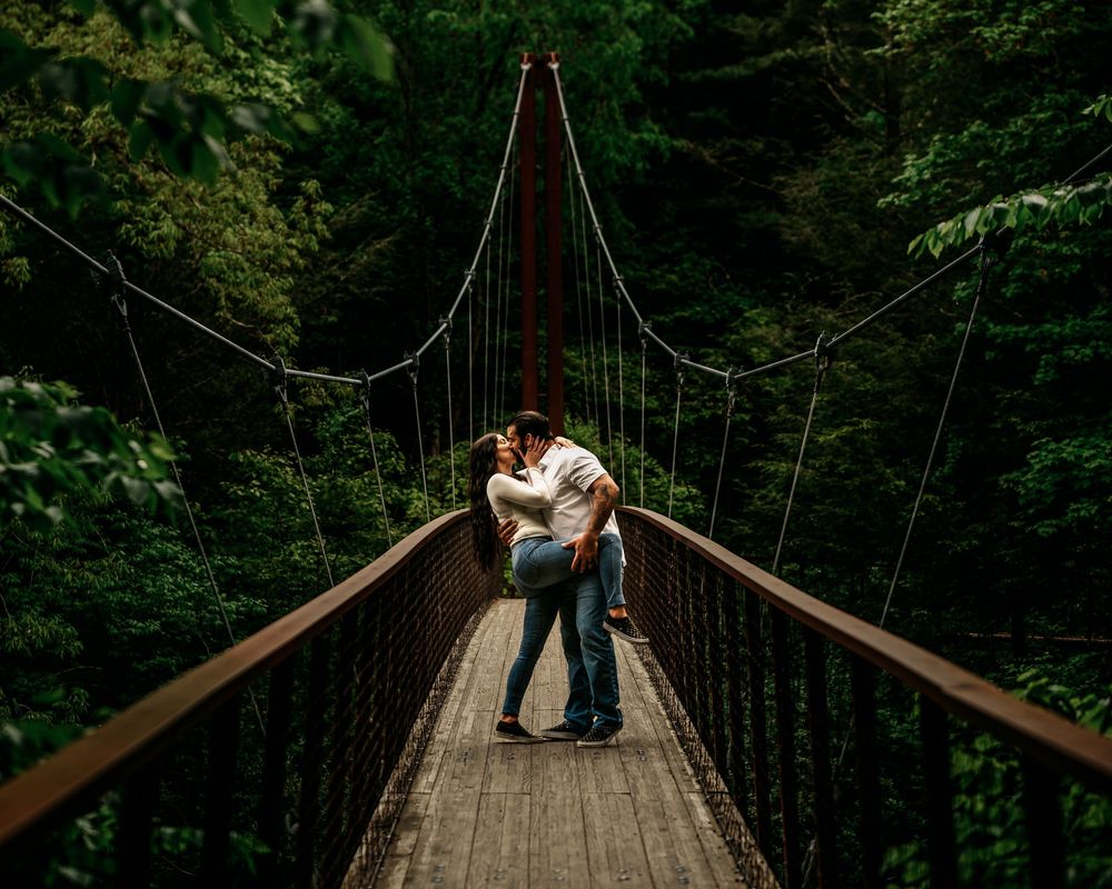 A romantic couple shares intimate moments on a wooden suspension bridge surrounded by lush green forest.
