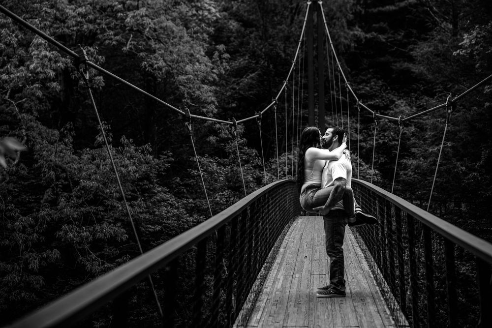 A person crosses a long suspension footbridge spanning through dense forest in black and white photography.