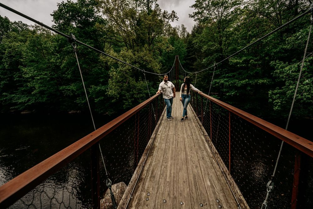 A rustic wooden suspension bridge stretches across a wooded ravine with people walking in the distance.