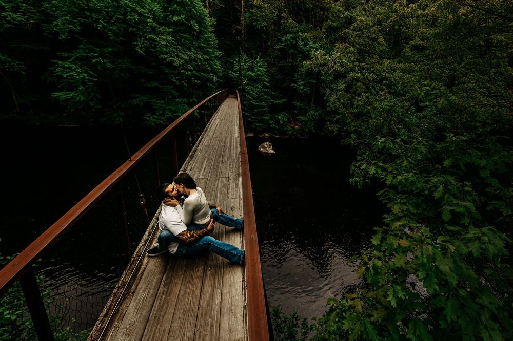 A wooden suspension bridge spans across a scenic forest river with lush green foliage surrounding it.