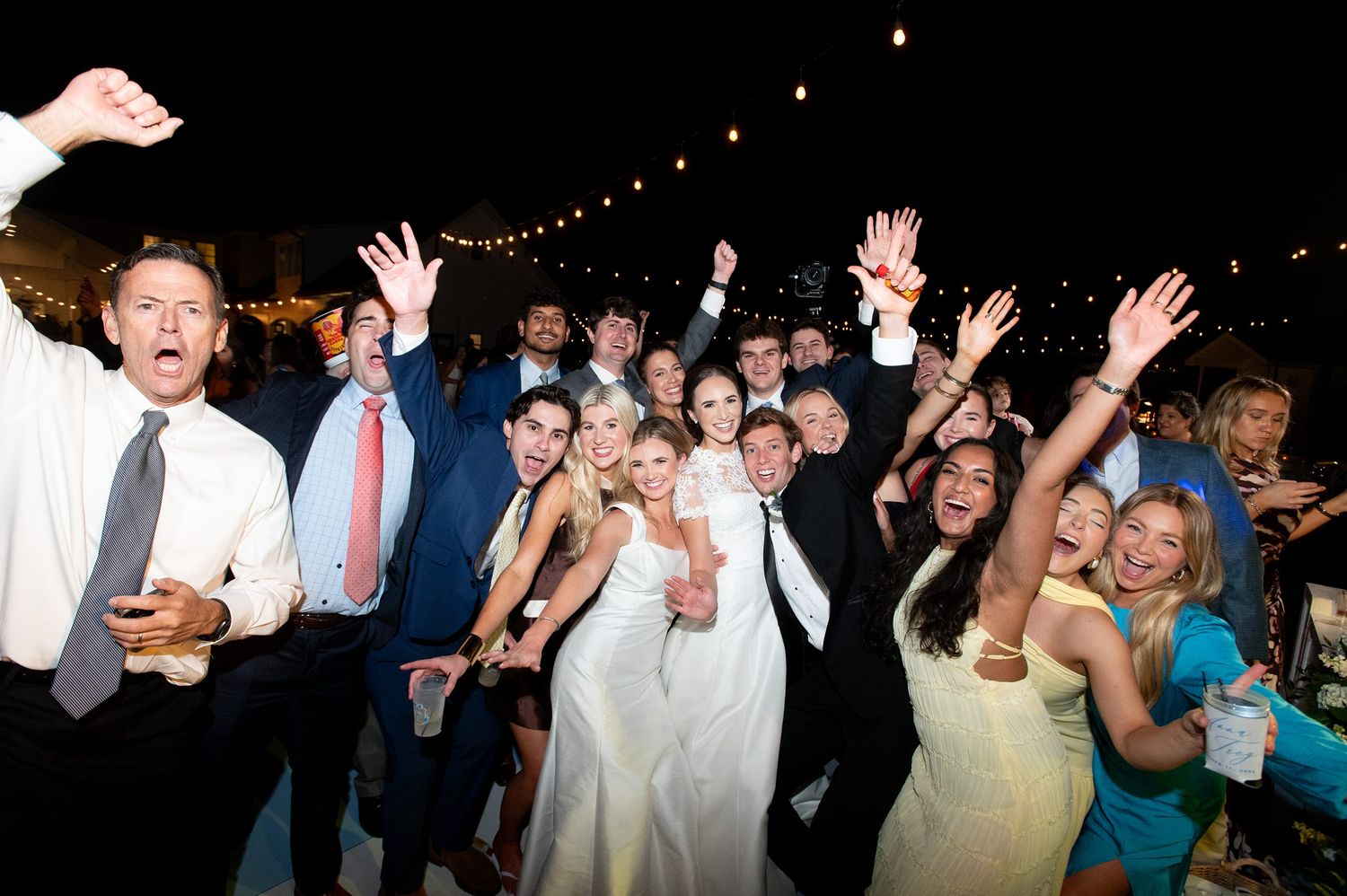 Wedding reception crowd dancing with hands in the air under string lights.