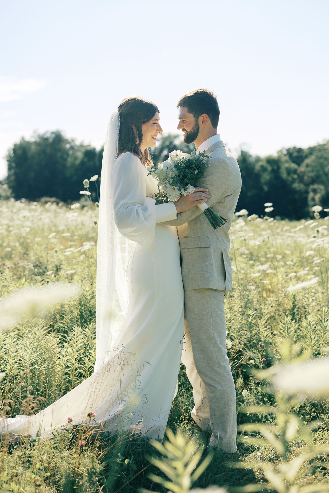 A couple shares an intimate moment in a sunlit wildflower meadow wearing light colored wedding attire.