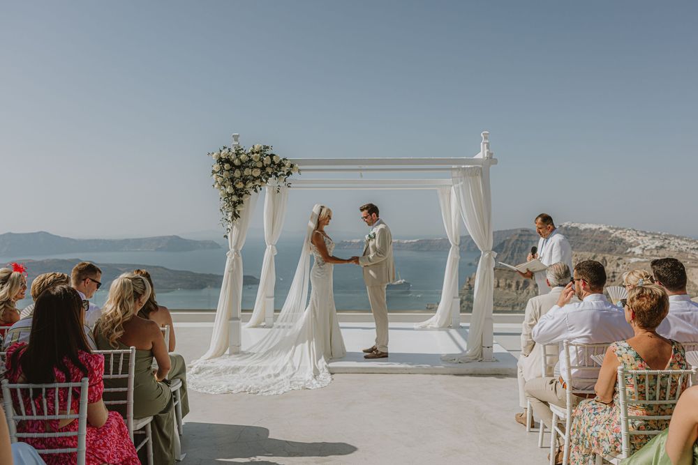 Oceanfront wedding ceremony setup with white drapes and chairs against blue sea views.
