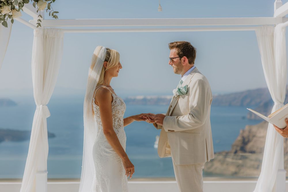 Wedding ceremony taking place under white draped arch overlooking Mediterranean coastal cliffs and sea views.