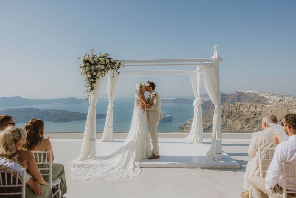 Outdoor wedding ceremony with white canopy arch decorated with flowers against stunning ocean backdrop.
