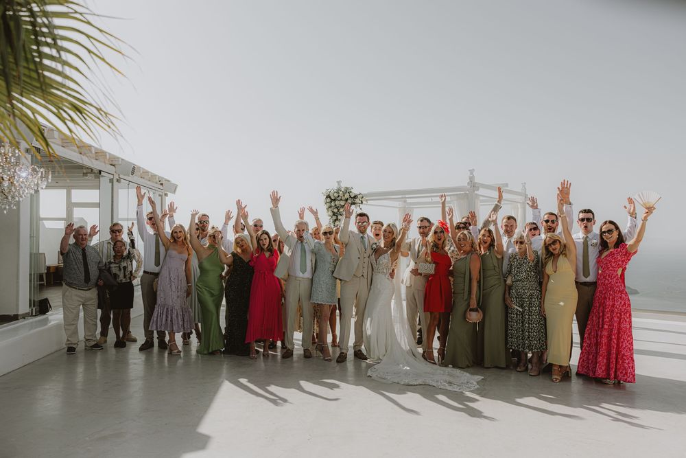 Large wedding party group photo with guests in colorful attire on white terrace overlooking ocean.