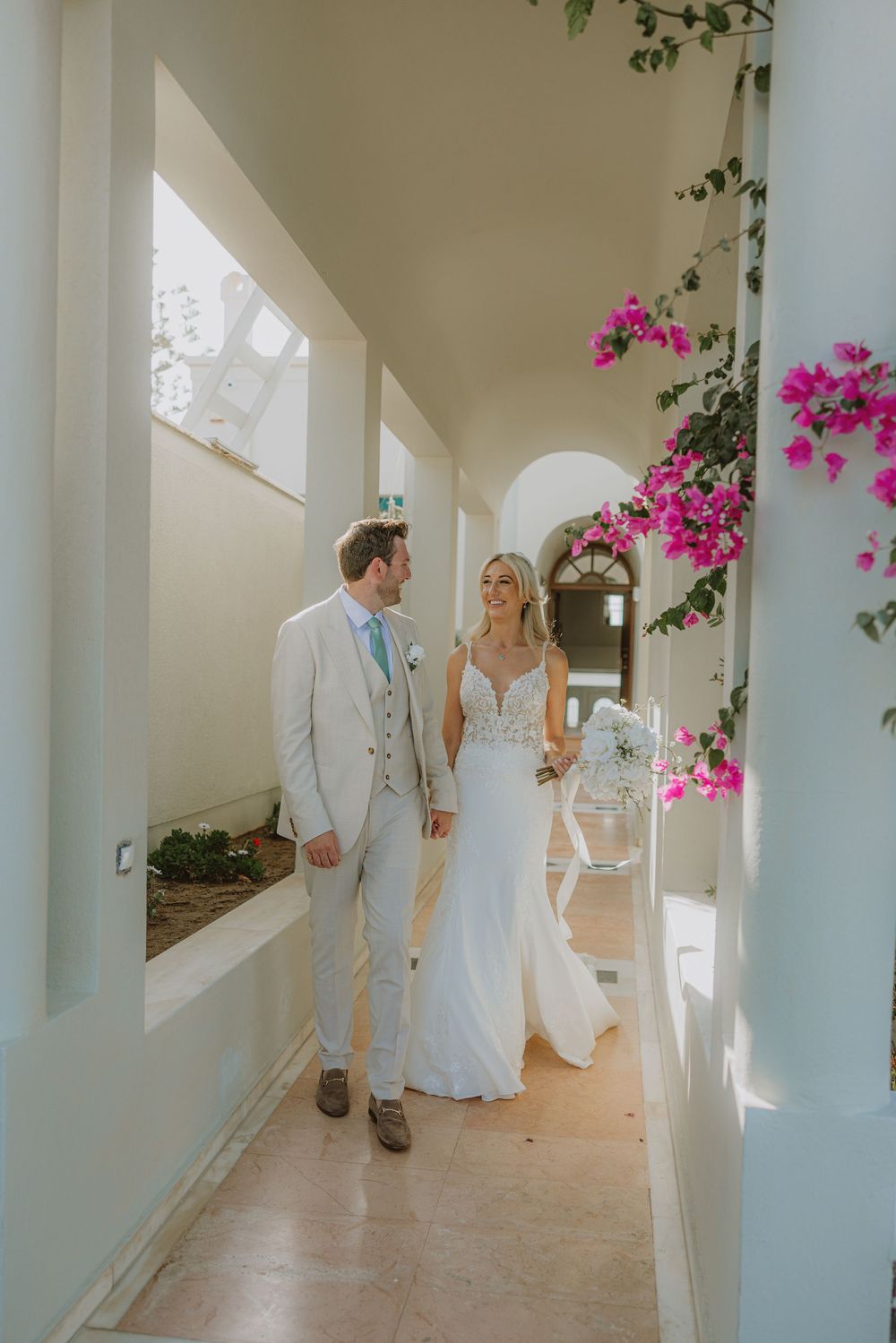 Wedding couple in elegant attire posing in white Mediterranean hallway with pink bougainvillea flowers.