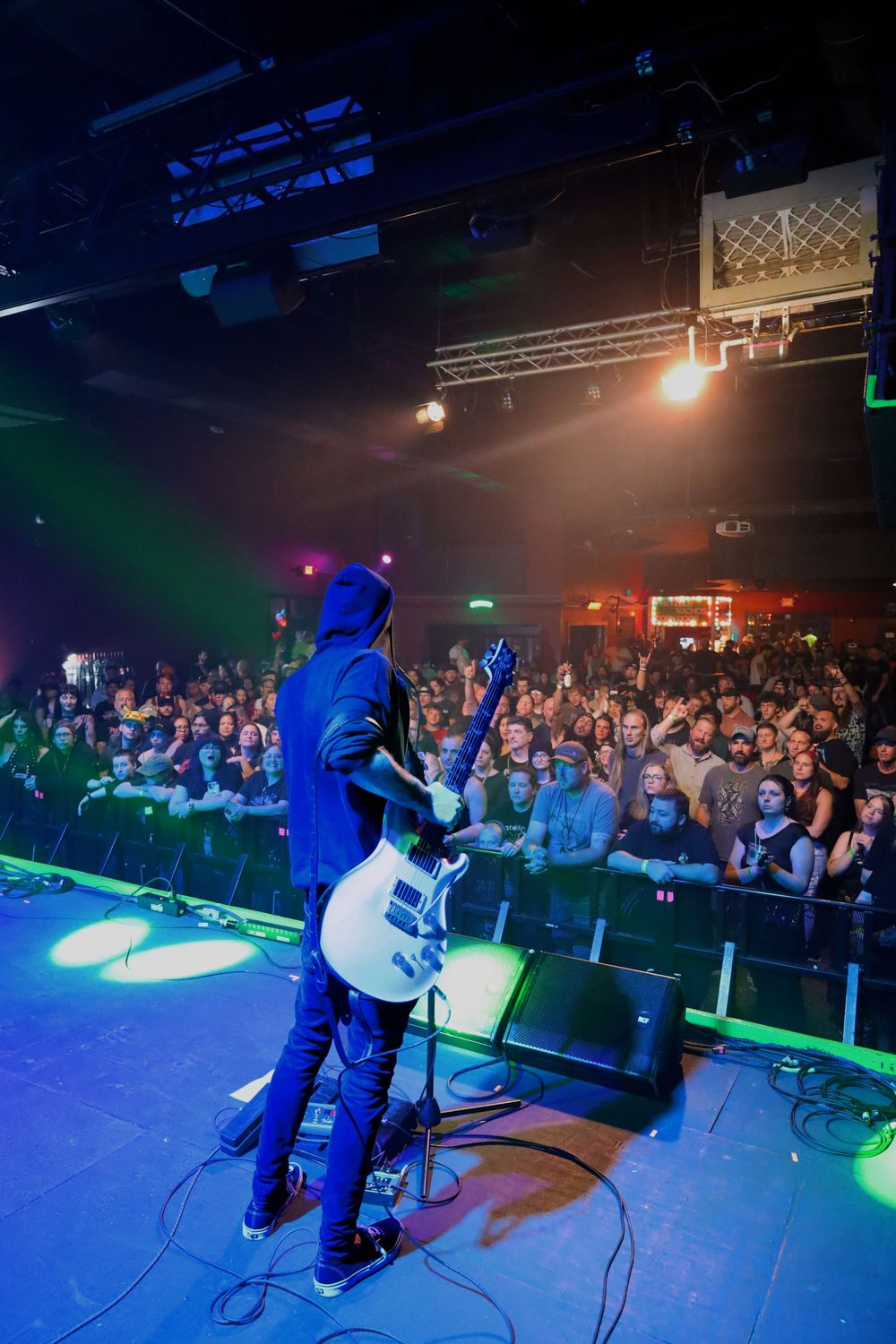 Performer in a dark hoodie on stage entertaining a large crowd at a nightclub with colorful lighting effects.