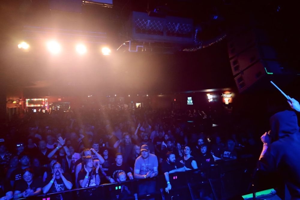 Concert crowd silhouettes with raised arms against blue stage lighting and atmospheric haze at a live music performance.