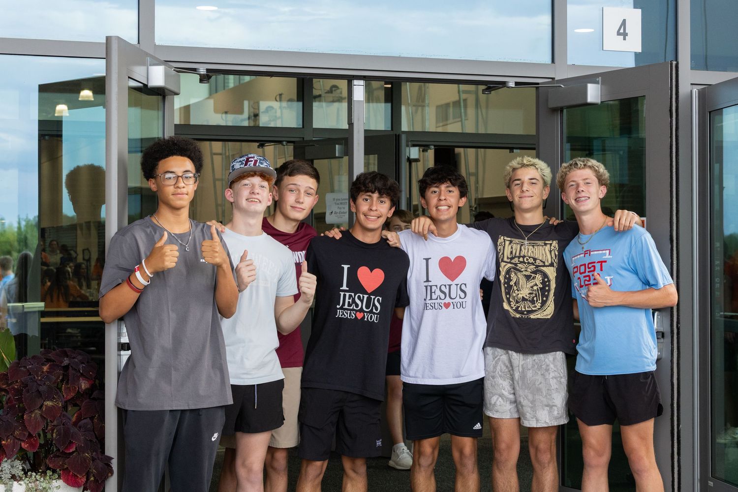 A group of students pose together in front of glass doors at a school entrance.